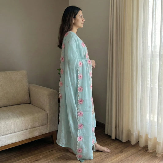 Woman wearing light blue kurti with pink floral embroidery, standing indoors by window
